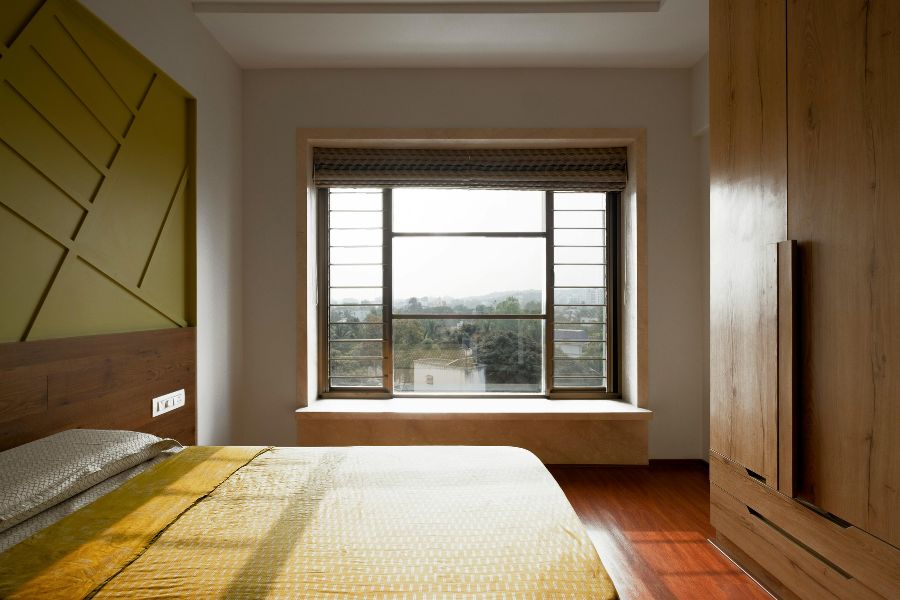 Chambre avec grande fenêtre à barreaux laissant entrer la lumière naturelle sur un lit jaune et un parquet en bois foncé.