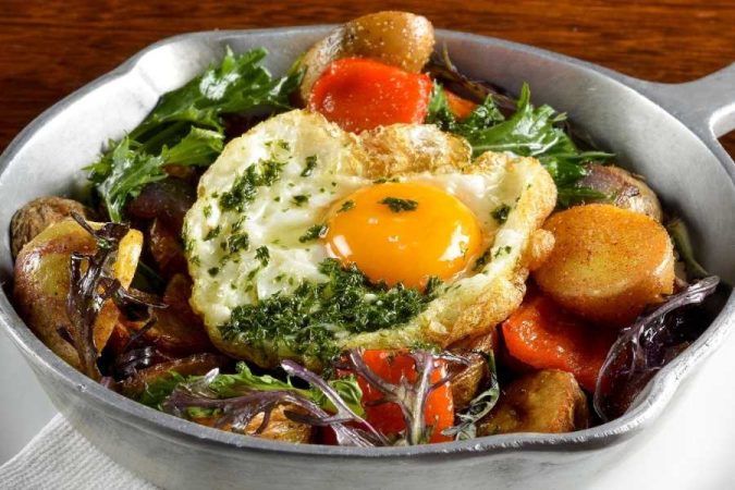 Poêle rustique garnie d'un œuf au plat aux herbes, pommes de terre rôties et légumes colorés, parfaite pour organiser un voyage gastronomique authentique.