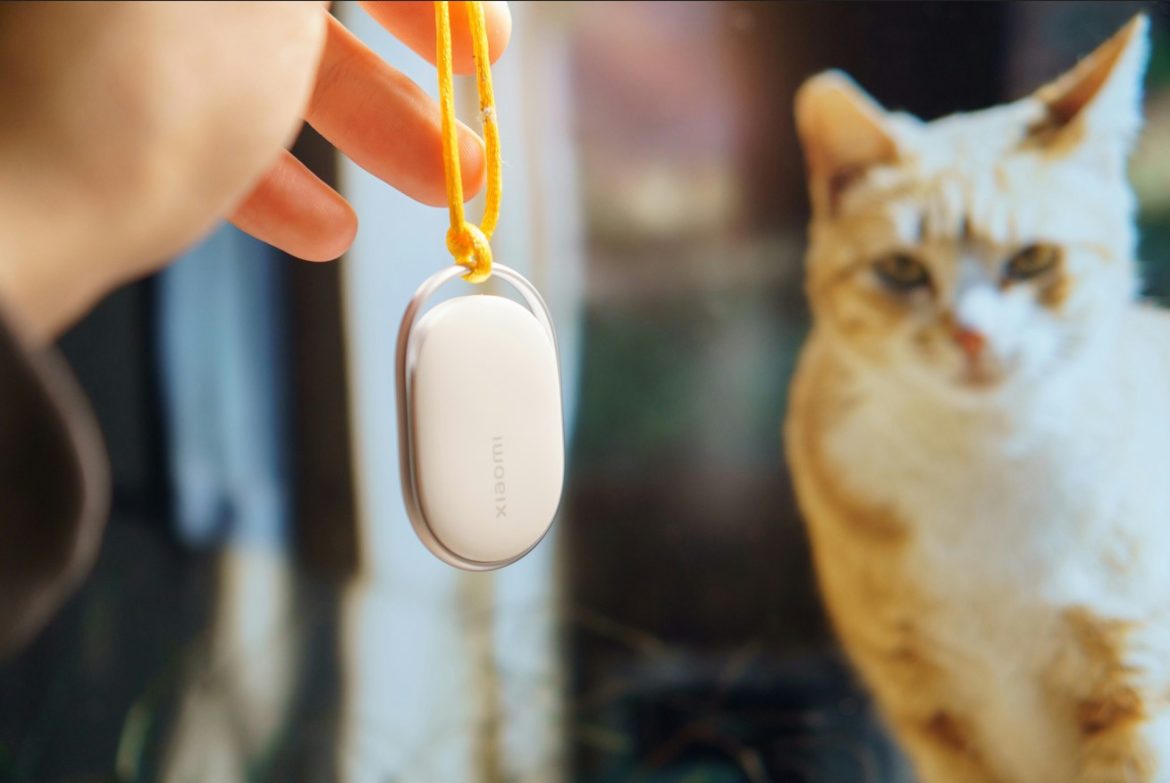 Une main tenant un traqueur connecté Xiaomi blanc avec un cordon jaune, devant un chat roux flouté en arrière-plan, illustrant l'IA et la Protection des Animaux.