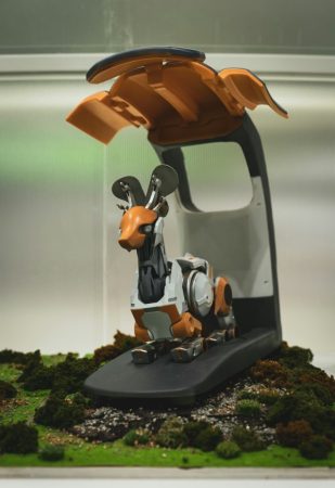 Maquette d'exposition d'un robot en forme de cerf orange et blanc, doté de capteurs, émergeant d'une capsule pour la surveillance faune intelligente sur un socle de mousse.