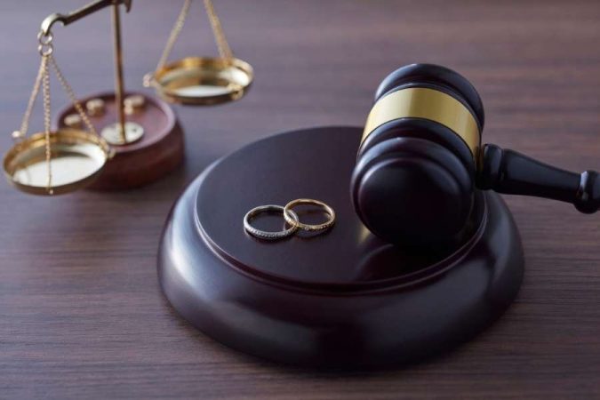 Un marteau de juge repose sur une table en bois sombre, avec deux alliances posées devant lui et une balance de justice en arrière-plan flou.