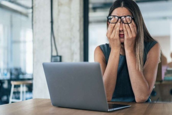 Une femme portant des lunettes et se frottant les yeux, assise devant un ordinateur ouvert, a un visage fatigué après une longue journée de travail.