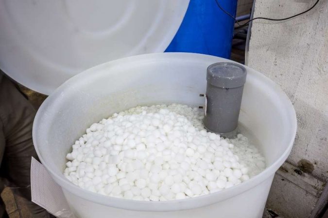 Un bac de stockage en plastique blanc, rempli à ras bord de pastilles de sel compactes et blanches, sert de réservoir à saumure pour un adoucisseur d'eau résidentiel.